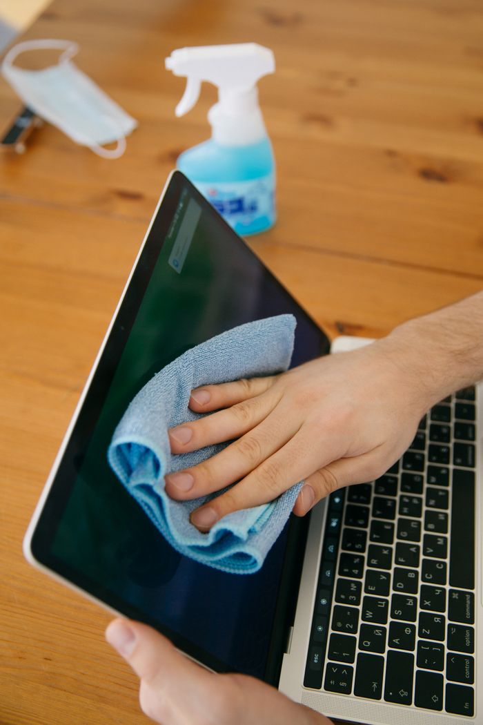 Une personne nettoie l’écran d’un ordinateur portable avec un chiffon bleu et un vaporisateur, posé sur une table en bois, pour l’hygiène.